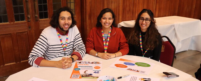 Three smiling students seated at a table with leaflets and pens